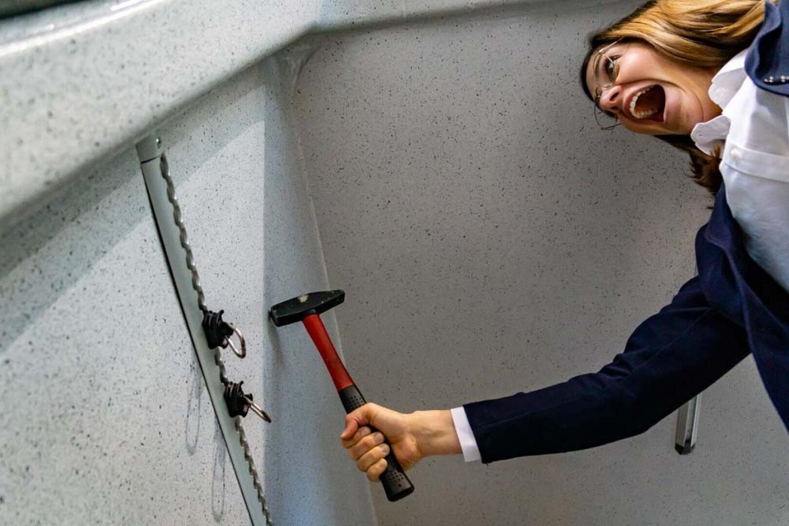 Eine frustriert wirkende Frau hält einen Hammer in die Nähe von Schlüsseln, die in einem Büro oder einem öffentlichen Raum an der Wand hängen, als ob sie sie zerschlagen wollte.