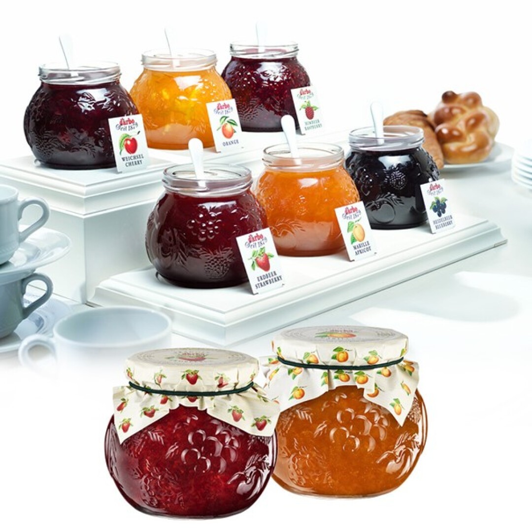 Sechs Gläser mit verschiedenen Fruchtmarmeladen, Etiketten und weißen Löffeln stehen auf weißen Ständern. Vorne sind zwei Gläser mit dekorativen Deckeln zu sehen. Im Hintergrund sind Teetassen und Gebäck zu sehen.