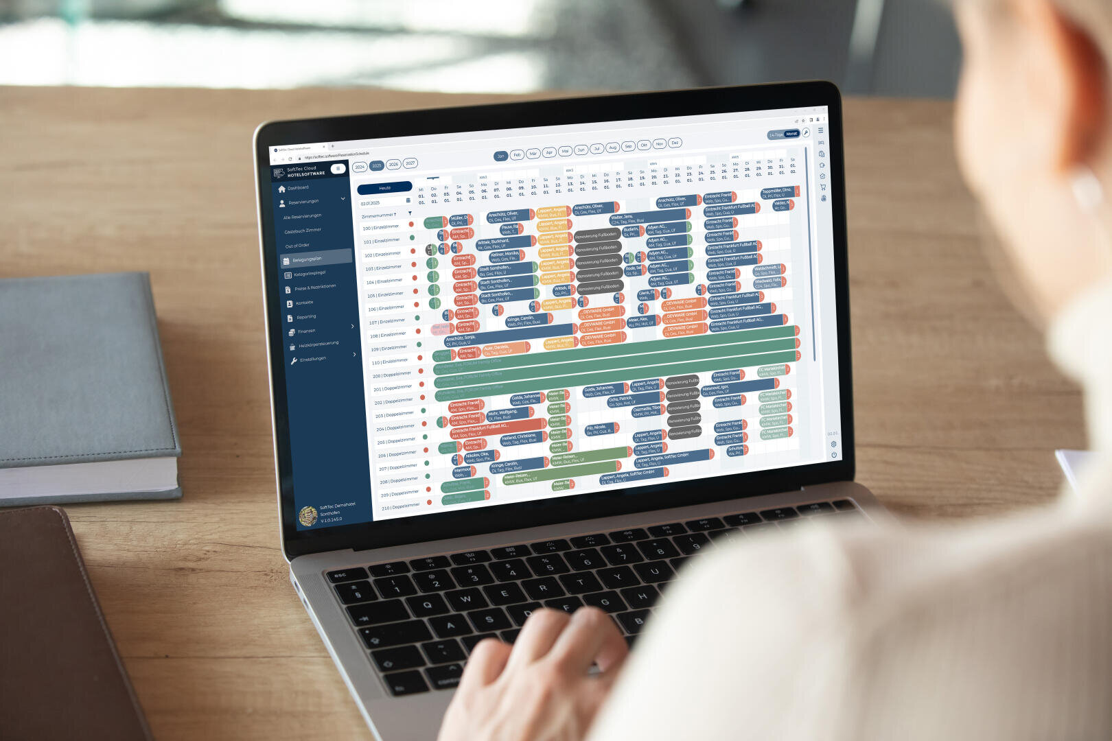 Eine Person nutzt einen Laptop, auf dem eine komplexe, farbkodierte Termin- oder Kalenderoberfläche mit mehreren Zeilen und Spalten angezeigt wird, möglicherweise für Projektmanagement oder Terminplanung. Im Hintergrund sind ein Holztisch und geschlossene Notizbücher zu sehen.
