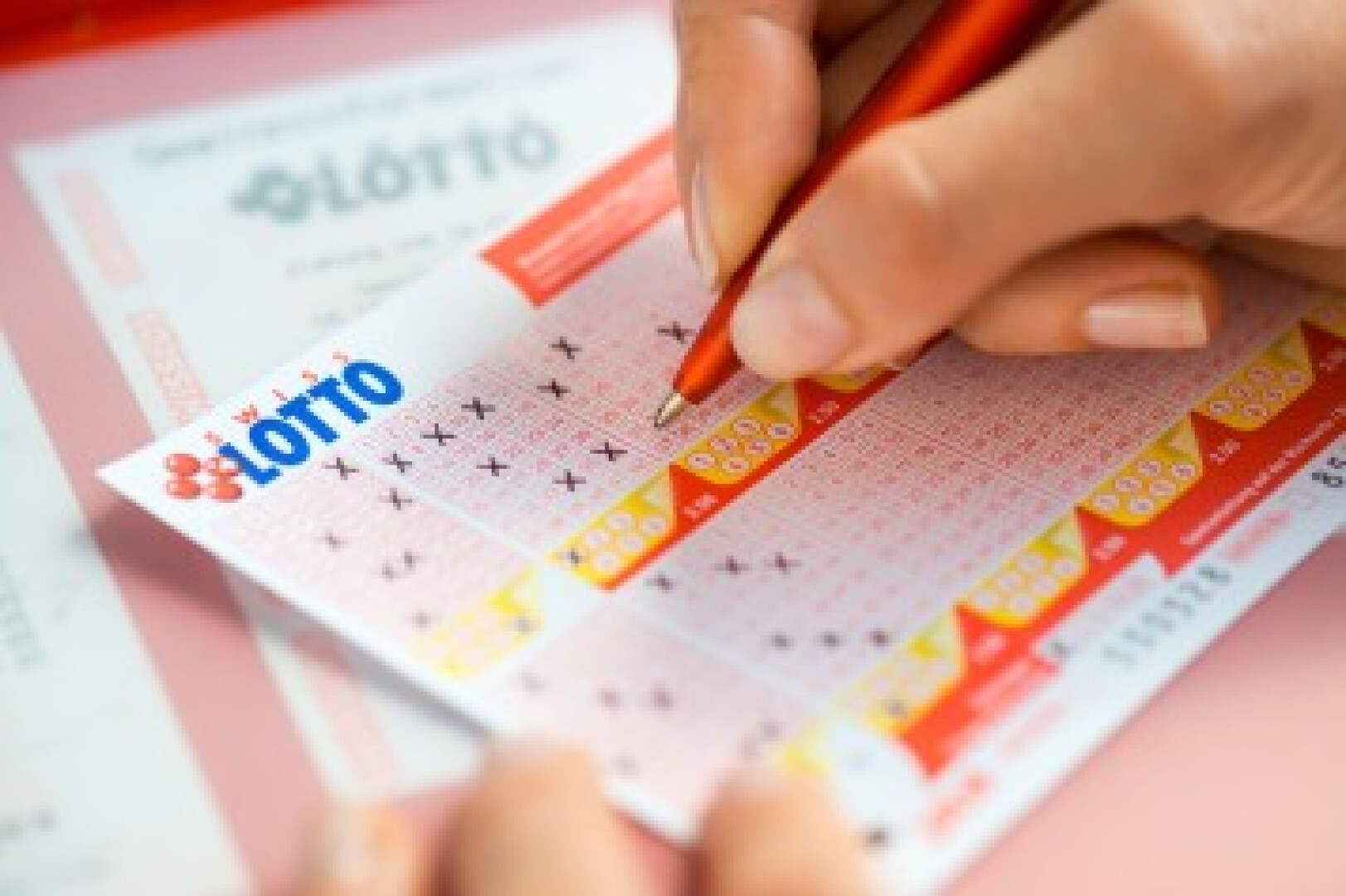 Eine Person markiert mit einem roten Stift Zahlen auf einem Lottoschein, wobei darunter teilweise ein weiterer Lottoschein sichtbar ist.