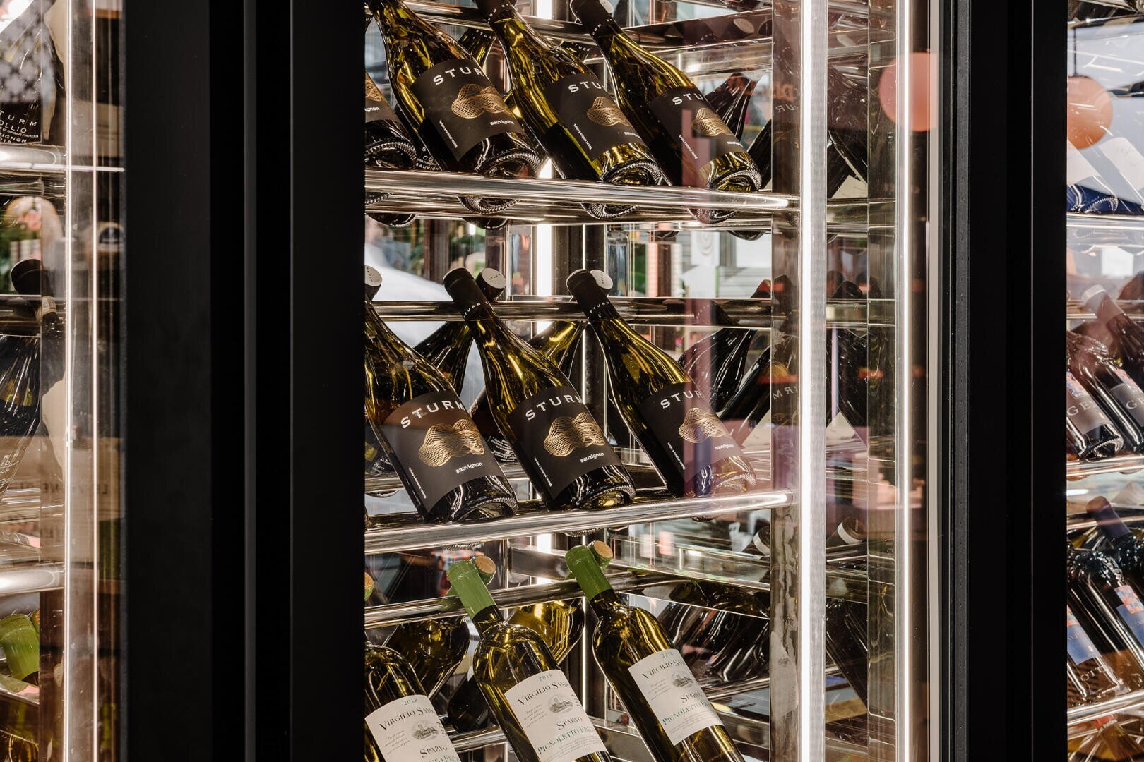 Weinflaschen sind auf schrägen Metallregalen im Inneren eines gläsernen Weinkühlschranks angeordnet, mit sichtbaren Etiketten auf mehreren Flaschen. Die Szene spiegelt eine moderne und hochwertige Umgebung für die Weinlagerung wider.