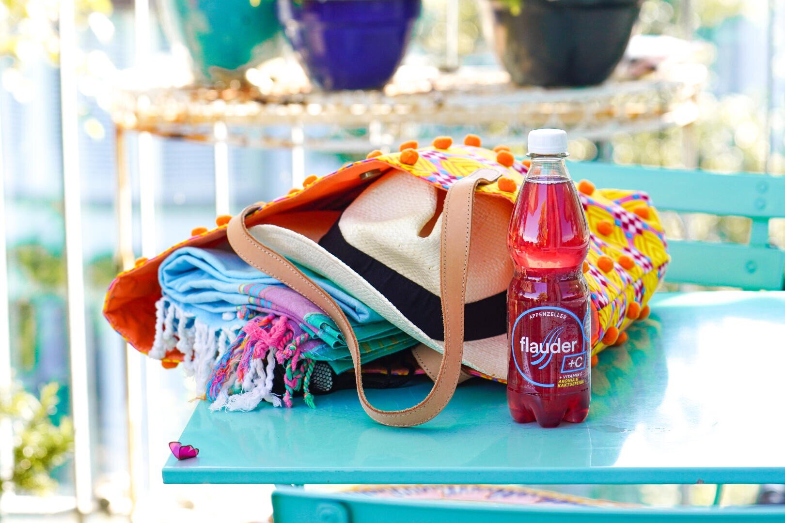 Eine bunte Strandtasche mit Handtüchern und einem Sonnenhut steht auf einem türkisfarbenen Tisch neben einer Flasche roter Flayder-Limonade, im Hintergrund stehen Topfpflanzen auf einem Regal.