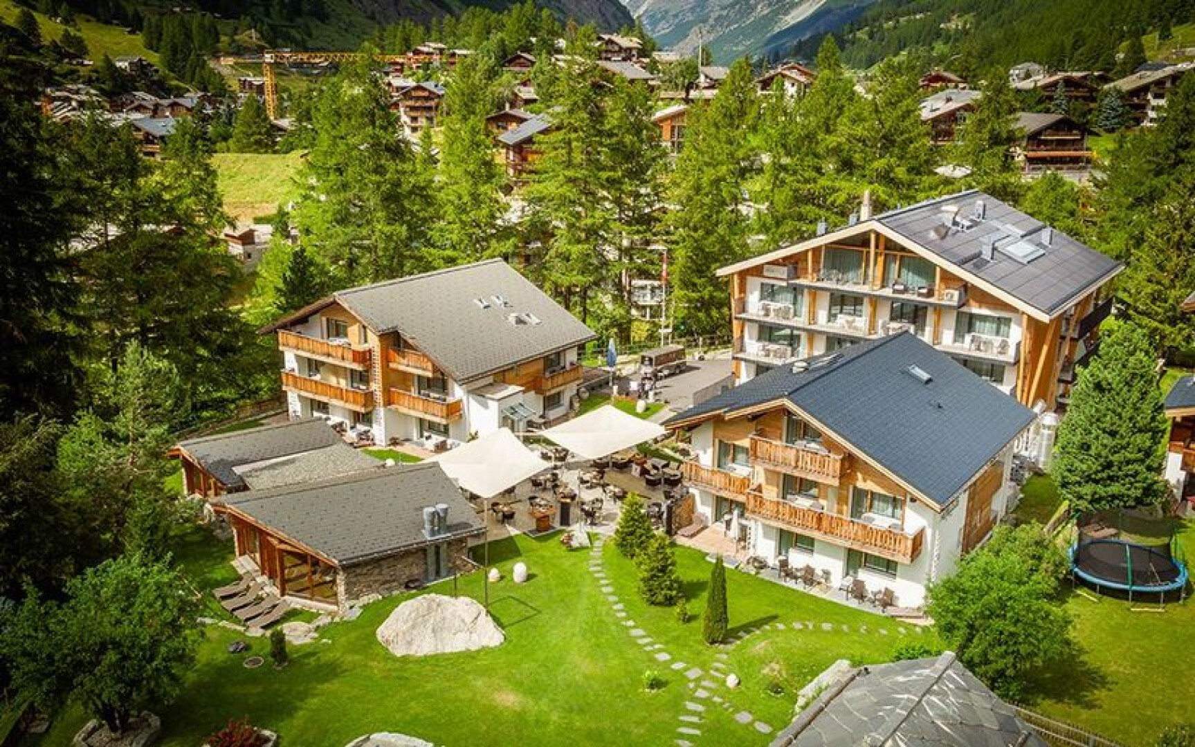 Luftaufnahme einer Gruppe moderner Holzchalets, umgeben von üppigen grünen Rasenflächen, Bäumen, Sitzbereichen im Freien und einer nahegelegenen Berglandschaft in einer malerischen Dorfkulisse.