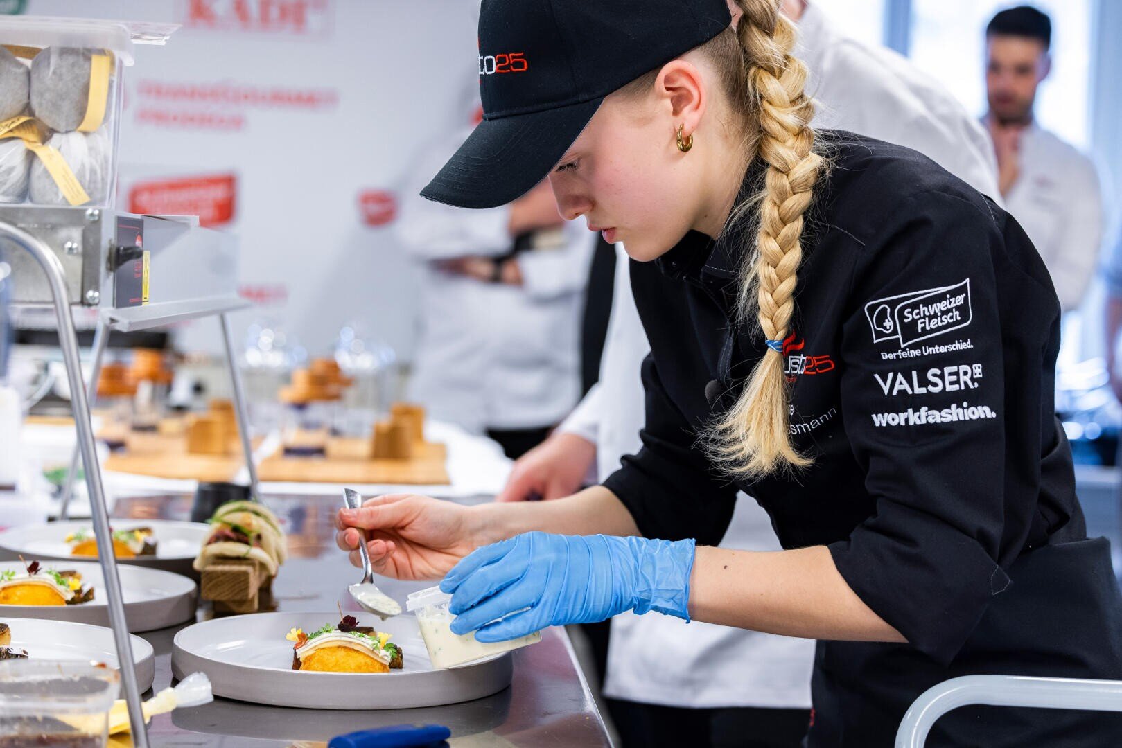 Ein Koch in schwarzer Uniform, mit Haube und blauen Handschuhen richtet in einer professionellen Küche sorgfältig Speisen an, während andere im Hintergrund zuschauen. Der Arbeitsbereich ist voller Geschirr und Küchenutensilien.