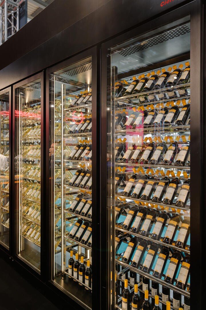 Ein großer verglaster Weinkühlschrank ist mit Reihen von passenden Weinflaschen gefüllt, die horizontal auf Metallregalen angeordnet sind, wobei die Reflexionen von Lichtern und Menschen auf den Glastüren sichtbar sind.