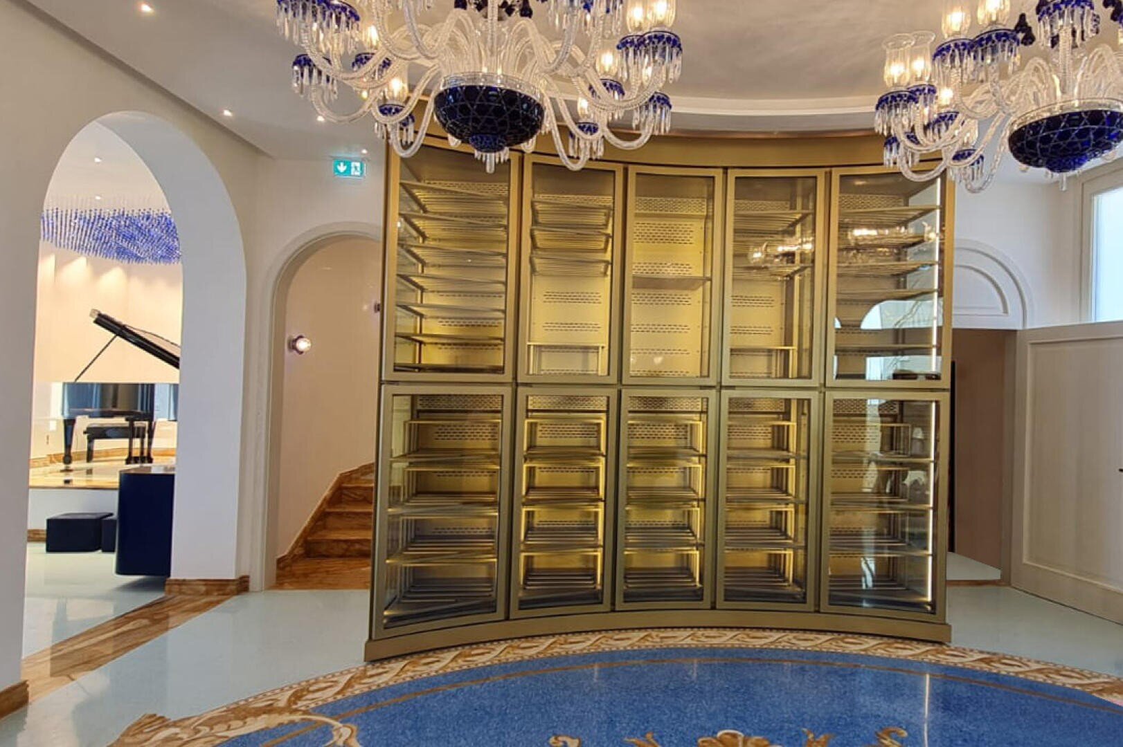 Ein luxuriöser Raum mit einem großen, geschwungenen, goldfarbenen Regal, verschnörkelten blauen und kristallenen Kronleuchtern, einem gemusterten Fußboden und einem Flügel, der in einem angrenzenden Raum durch einen Torbogen sichtbar ist.