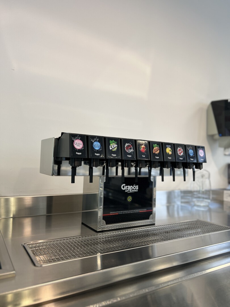 Ein Selbstbedienungs-Sodabrunnenautomat mit acht Getränkeoptionen, darunter Cola, Zitrone-Limette, Orange und andere Limonaden, steht auf einer Edelstahltheke in einem sauberen, modernen Ambiente.