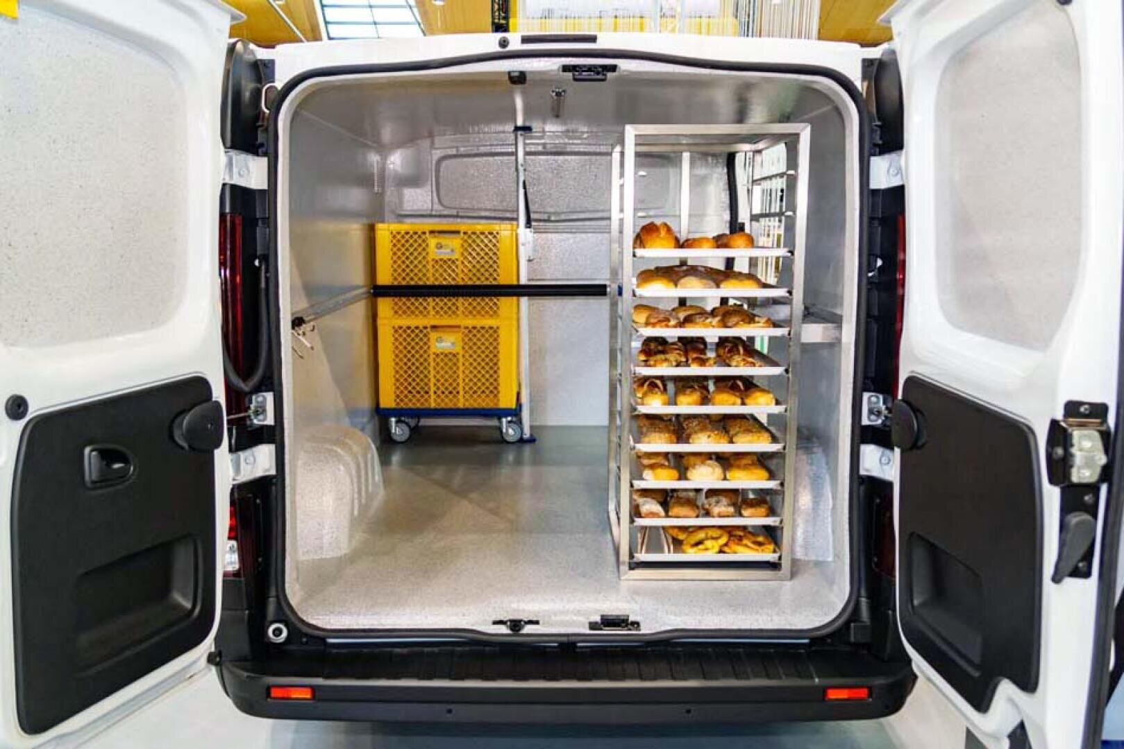 Das Bild zeigt die offenen Hintertüren eines gekühlten Lieferwagens. Im Inneren befinden sich zwei gelbe Kisten und ein Metallgestell, die mit Backwaren oder Brot gefüllt und für den Transport geordnet sind.