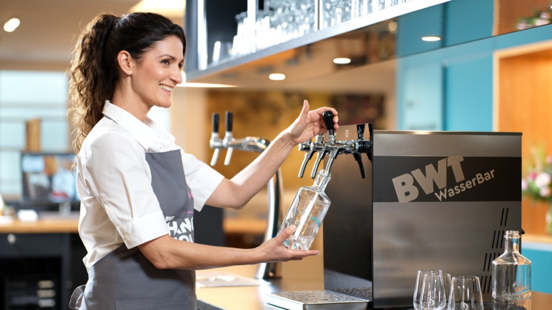 Une femme portant un tablier gris sourit en remplissant une bouteille en verre d'eau provenant d'un distributeur BWT WasserBar sur un comptoir moderne, avec des verres et d'autres bouteilles à proximité.