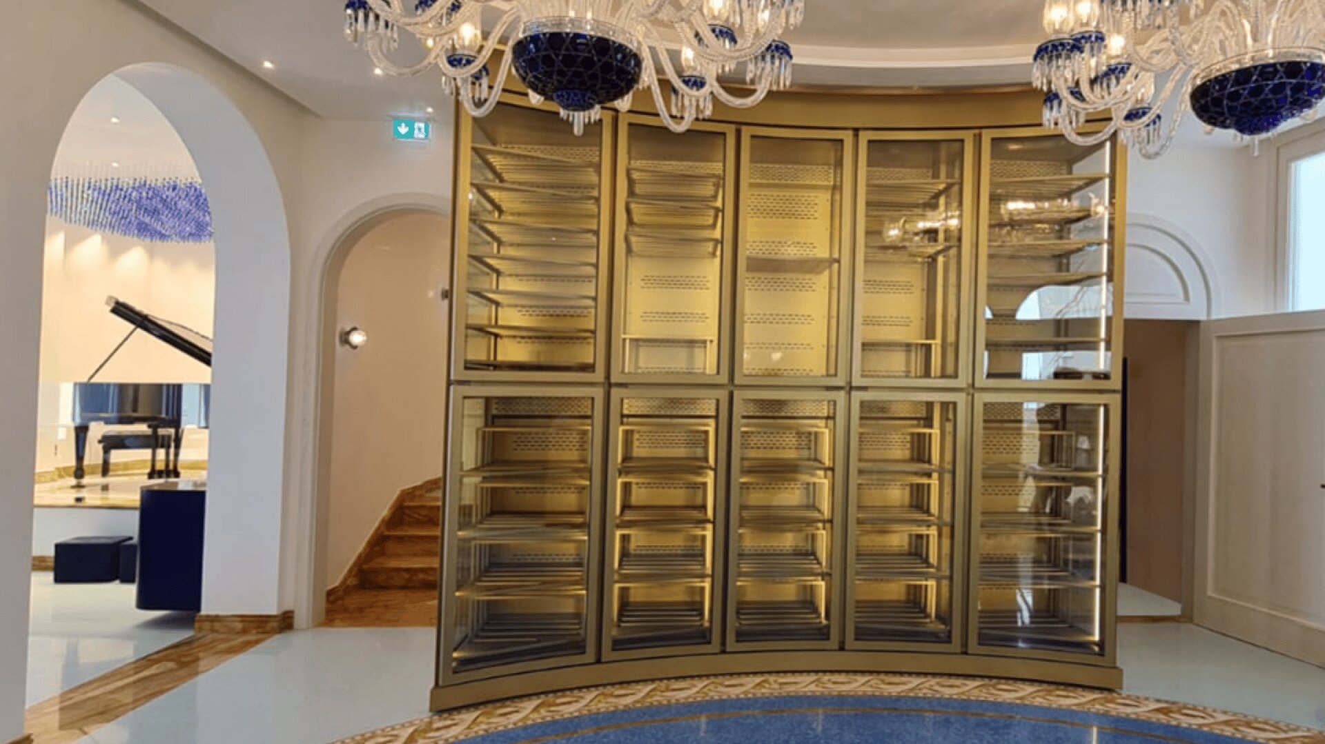 Une grande et élégante vitrine dorée aux étagères vides se trouve dans une pièce luxueuse avec des lustres, un sol en marbre et un piano à queue visible à l'arrière-plan.