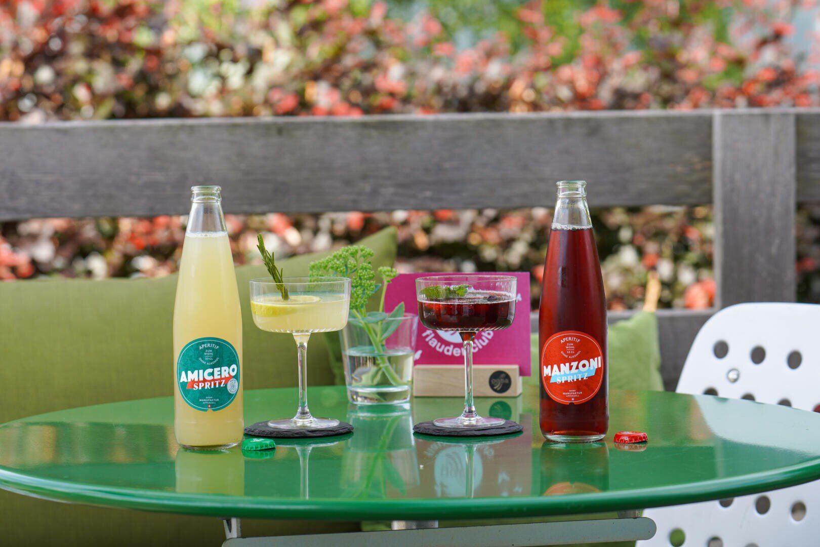 Zwei Glasflaschen und zwei Cocktailgläser mit bunten Getränken stehen auf einem grünen Gartentisch. Im Hintergrund sind Blumen und ein unscharfes Holzgeländer zu sehen. Ein Glas Wasser und eine Speisekarte sind ebenfalls zu sehen.