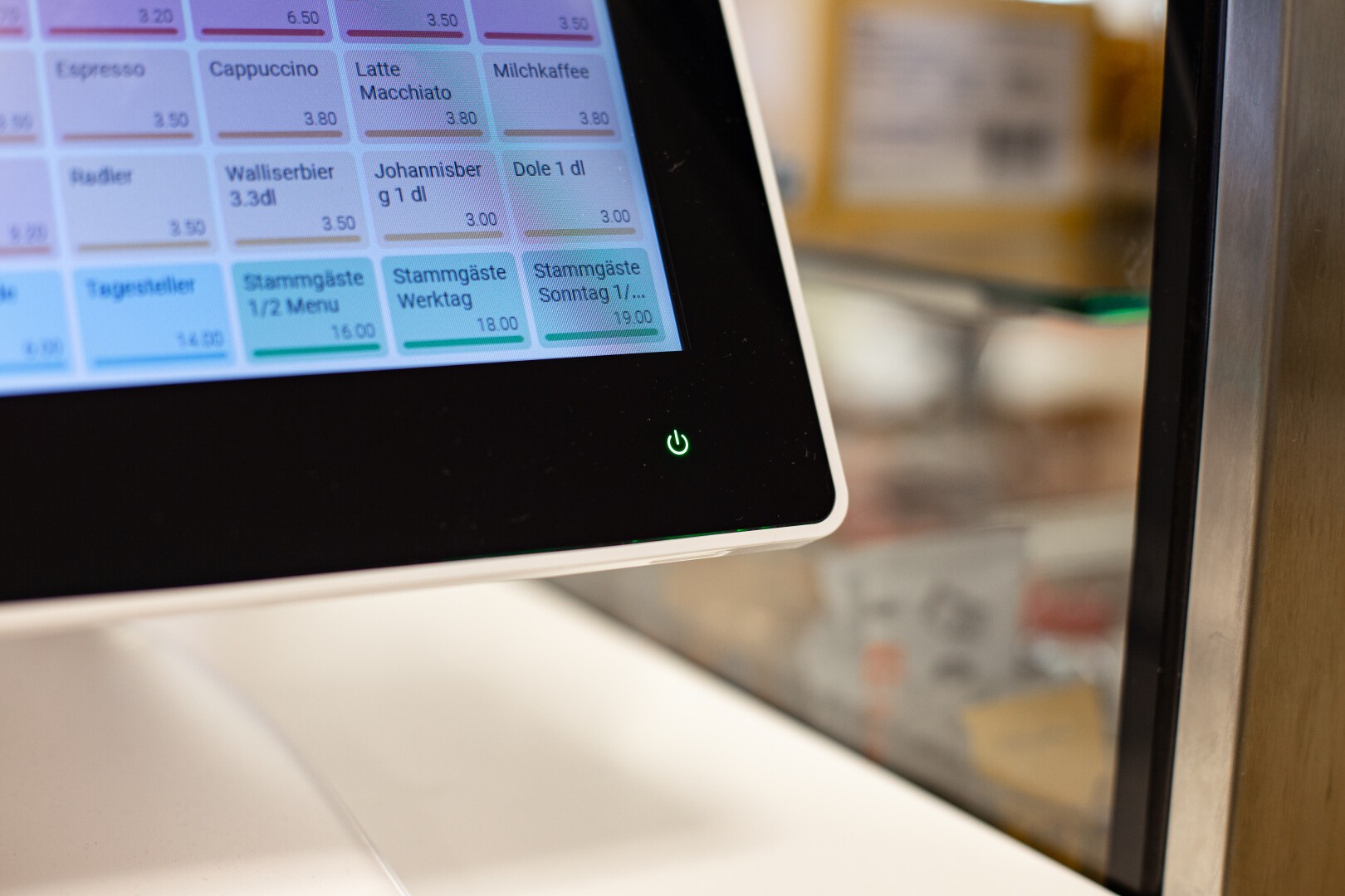 Nahaufnahme eines Touchscreen-Bestellsystems mit einer Speisekarte mit verschiedenen Getränken und Preisen auf Deutsch, neben einem grünen Einschaltknopf auf dem Gerät. Der Hintergrund ist unscharf.
