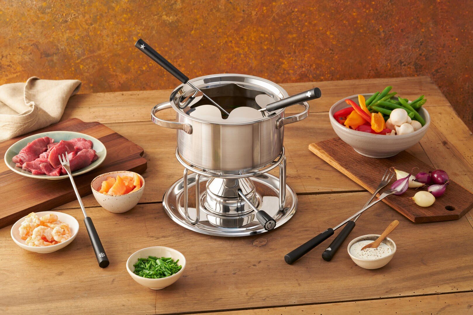 Ein Fonduetopf aus Edelstahl mit Gabeln steht auf einem Holztisch, umgeben von Schüsseln und Tellern mit rohem Fleisch, Garnelen, Gemüse, Soße und gehackten Kräutern, bereit für ein Fondue-Essen.