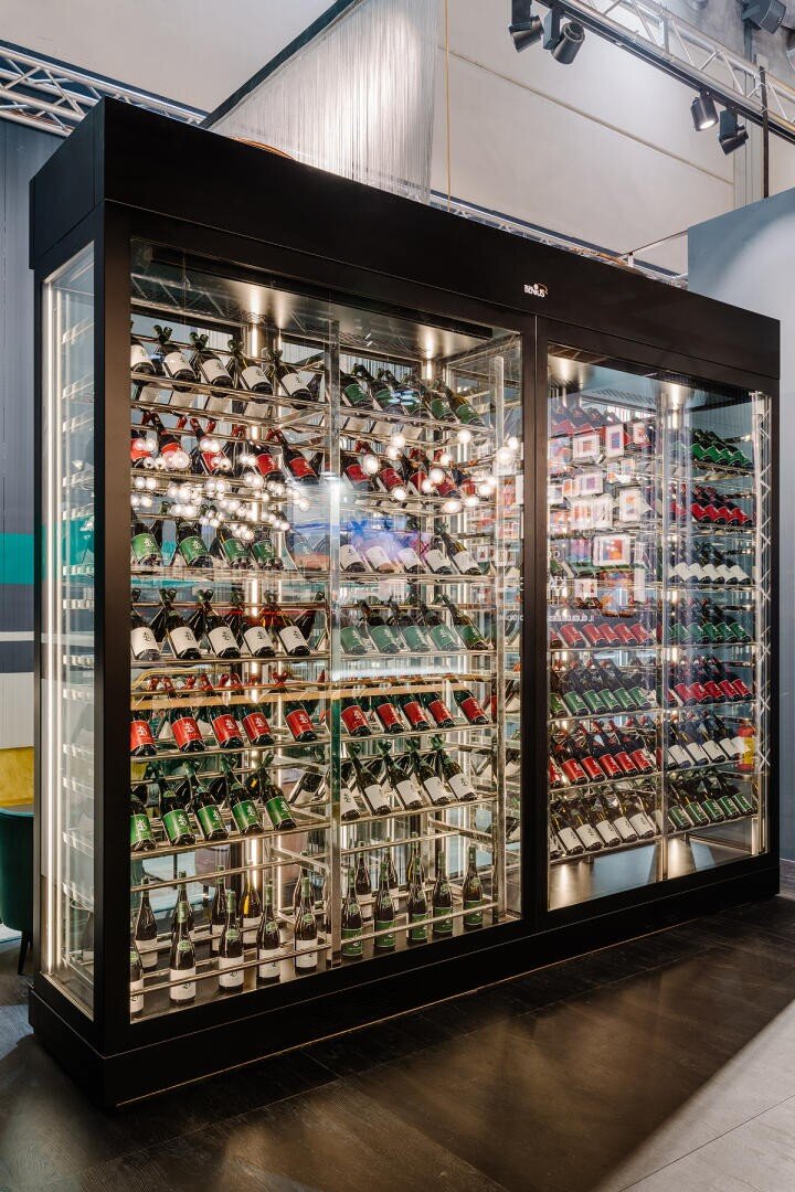 Ein moderner Weinkühlschrank aus Glas mit Metallregalen, gefüllt mit Rot-, Weiß- und Schaumweinflaschen, präsentiert in einem gut beleuchteten zeitgenössischen Rahmen.