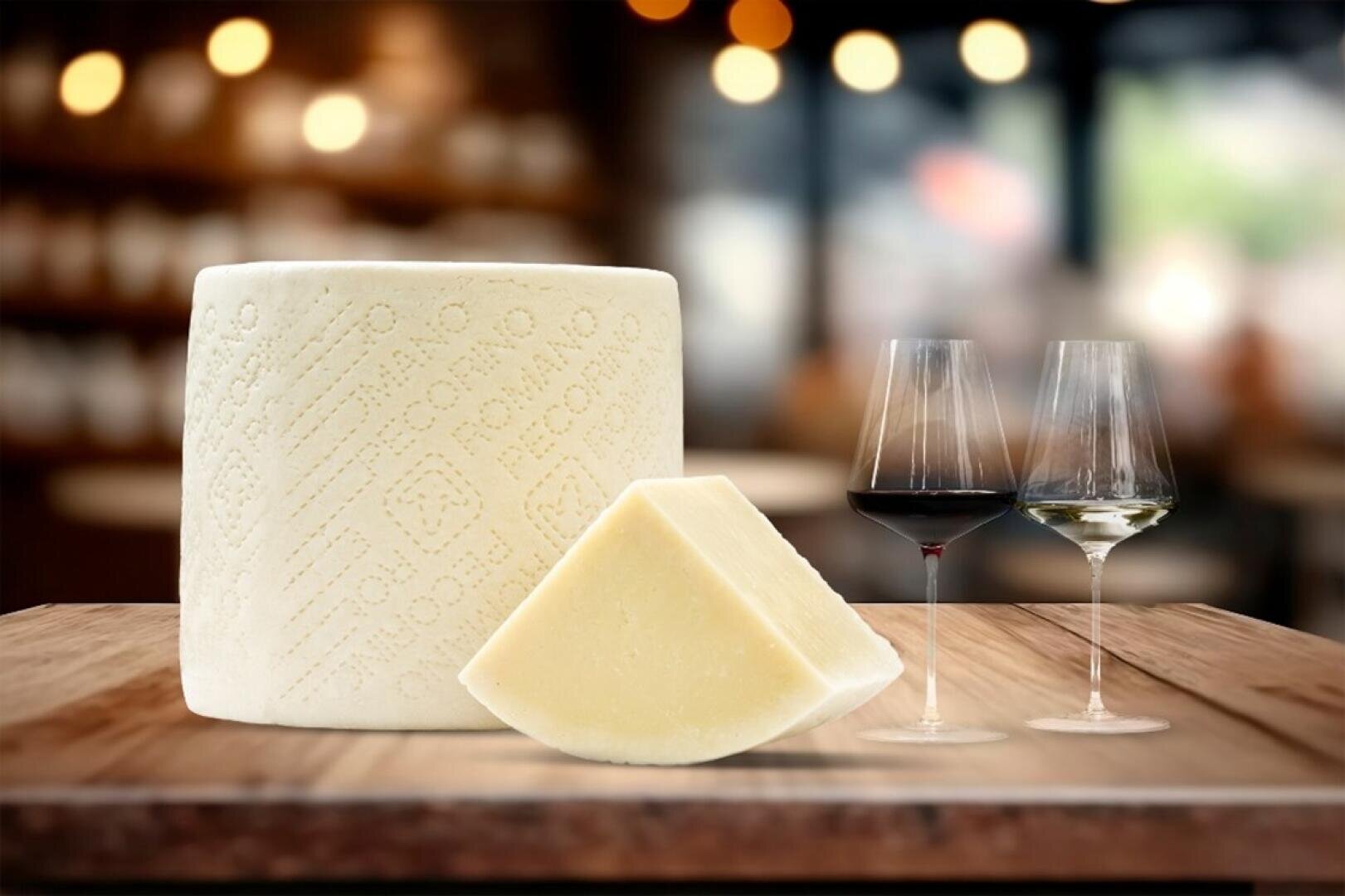 Ein Laib Schafskäse (Pecorino Romano) mit einem ausgeschnittenen Stück liegt auf einer hölzernen Unterlage neben zwei Gläsern Wein, einem roten und einem weißen, mit einem sanft verschwommenen Restaurant-Hintergrund.