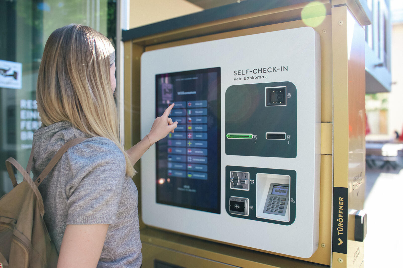 Eine Frau mit blonden Haaren und Rucksack nutzt einen Self-Check-in-Automaten mit Touchscreen. Der Automat zeigt verschiedene Optionen an und verfügt über Kartenfächer und ein Tastenfeld.