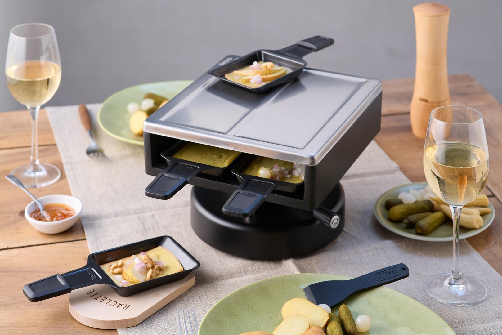 Ein Tisch-Raclette-Grill mit kleinen Schalen mit geschmolzenem Käse und Belag, umgeben von Tellern mit Kartoffelscheiben, eingelegten Gurken, Gläsern mit Weißwein und Besteck auf einem Holztisch.
