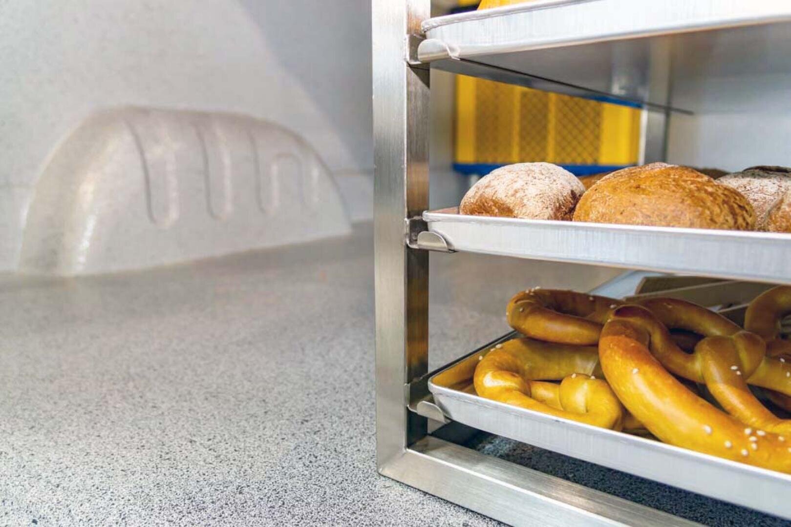 Drei Metallschalen mit Backwaren, darunter Brötchen und Brezeln, sind auf einem Gestell in einer Küche oder Bäckerei mit einer strukturierten Arbeitsplatte und einem industriellen Hintergrund gestapelt.