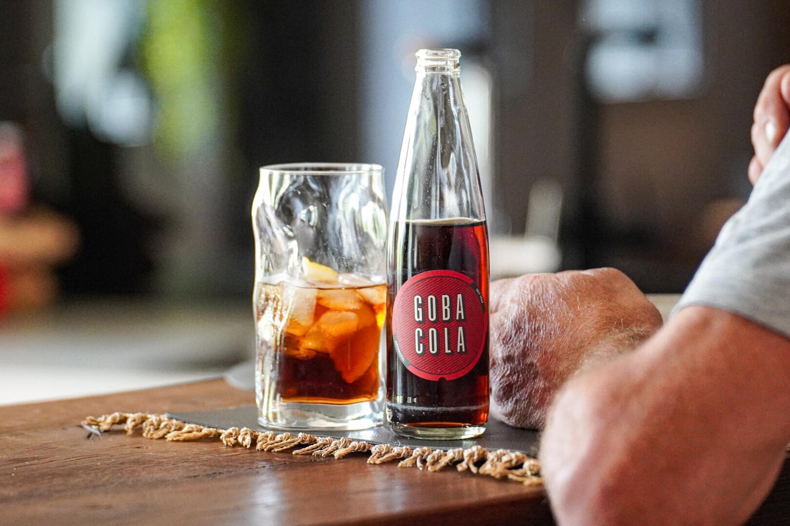 Eine Glasflasche mit der Aufschrift „Goba Cola“ steht neben einem mit Eis und Cola gefüllten Glas auf einem Holztisch. Der Arm einer Person ruht in der Nähe. Die Szene wirkt entspannt und ungezwungen in einem Innenbereich.