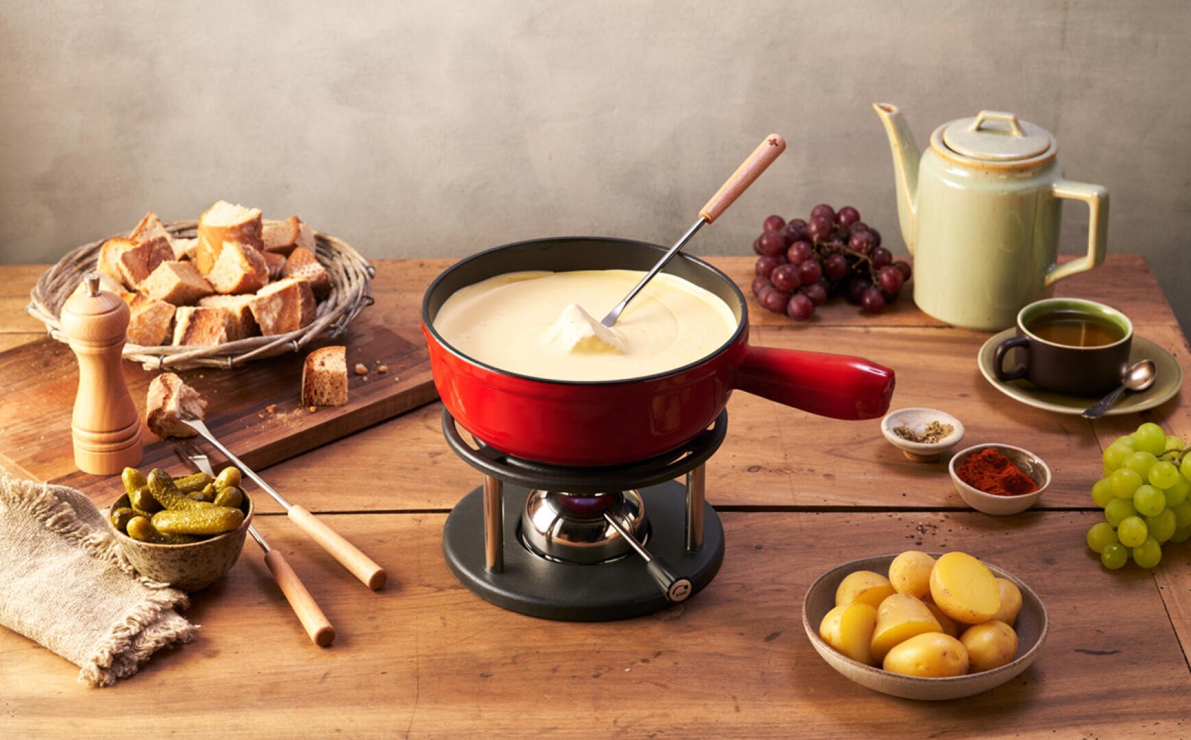 Ein roter Topf mit Käsefondue steht auf einem Brenner, umgeben von Brotwürfeln, Weintrauben, Salzkartoffeln, Essiggurken, Gewürzen und einer Teekanne mit einer Tasse Tee, alles auf einem Holztisch angeordnet.