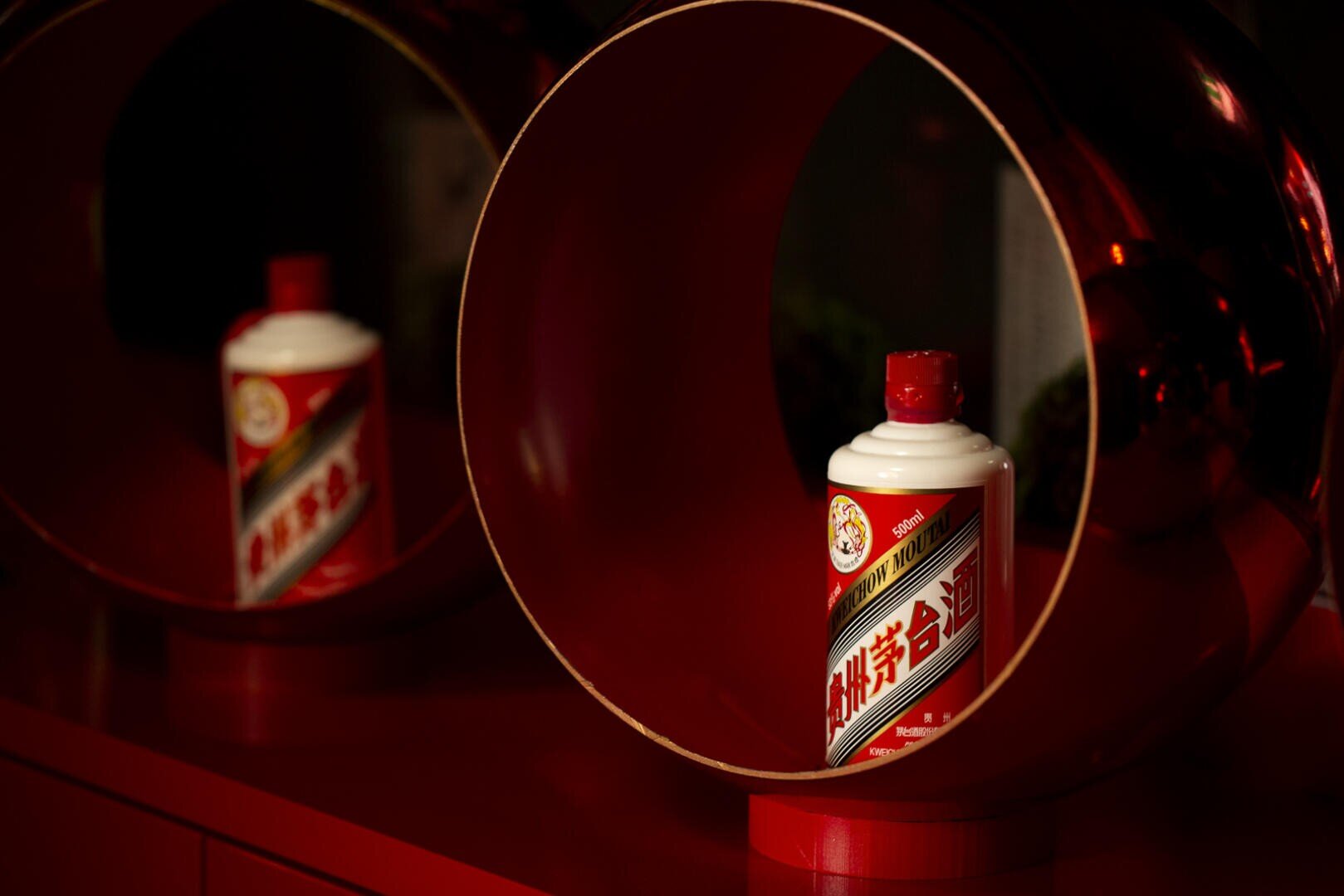 Eine Flasche Moutai-Likör mit rot-weißem Etikett steht in einem runden roten Metallrahmen. Im Hintergrund ist eine weitere identische Flasche zu sehen. Die Szene ist mit warmem Licht beleuchtet.