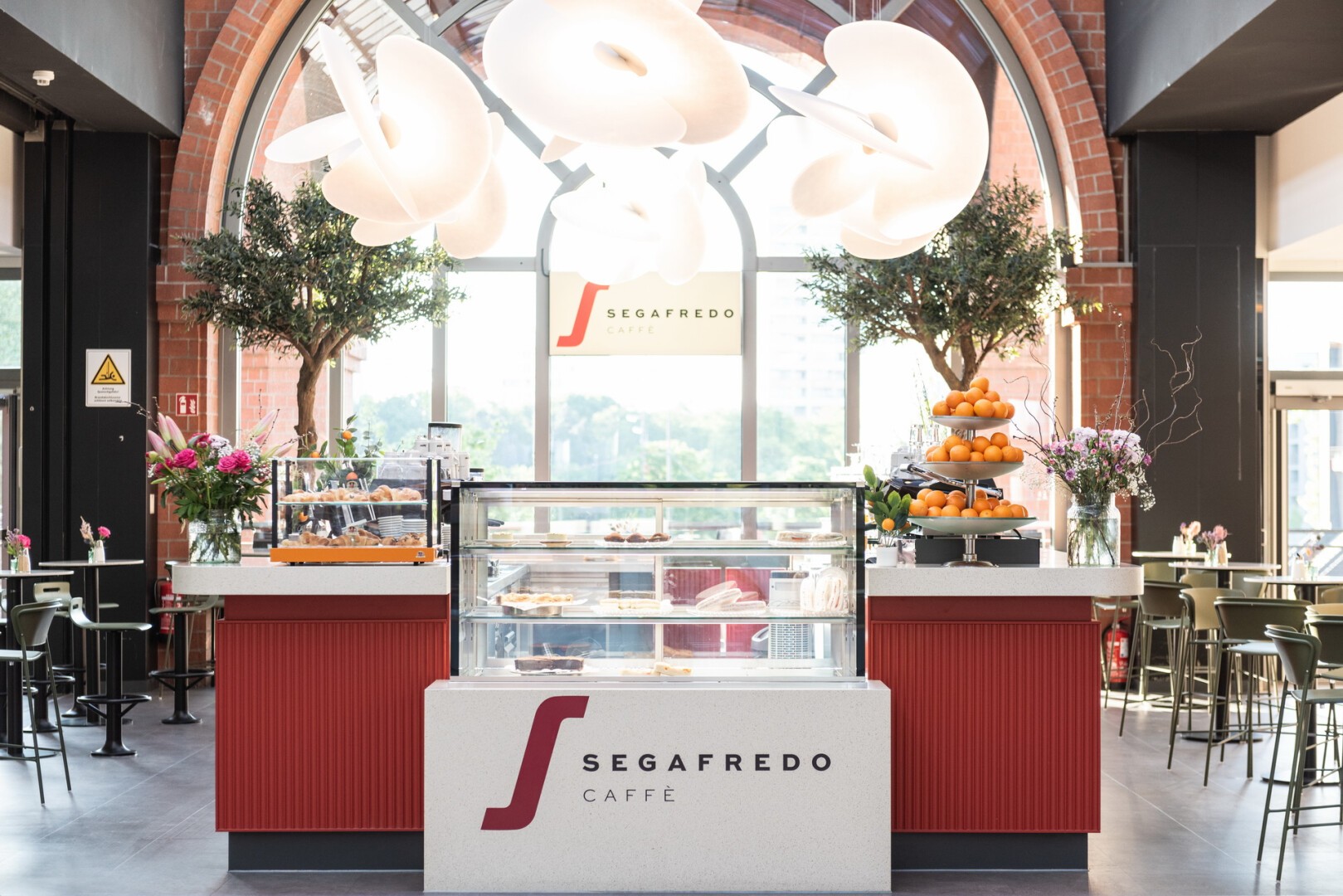 Eine moderne Segafredo Caffè-Theke mit Glasvitrinen voller Gebäck, gestapelten Orangen, Blumenarrangements und großen weißen Hängelampen, in einem hellen Café mit hohen Fenstern und Bäumen im Innenbereich.