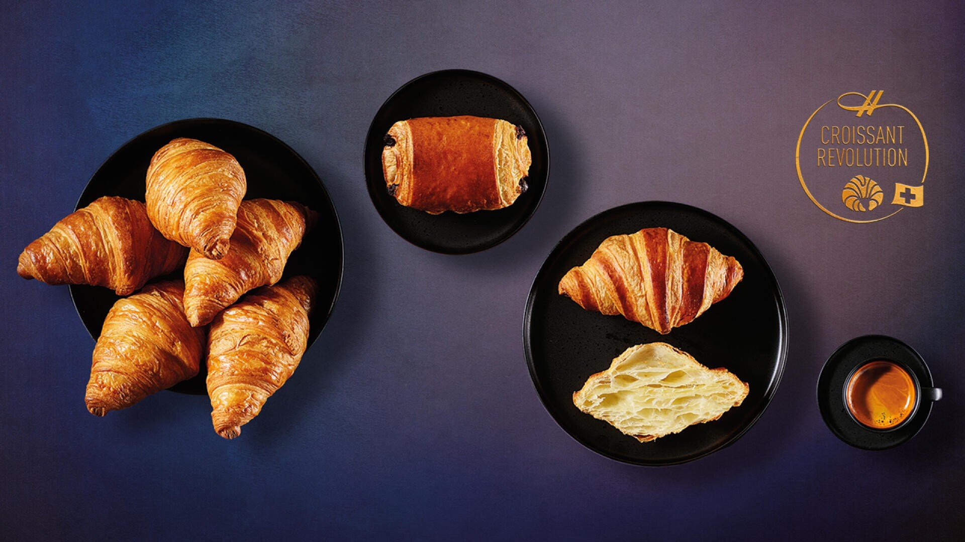 Eine dunkle Tischplatte mit einem Teller mit Croissants, einem Teller mit einem Pain au Chocolat, einem Teller mit einem ganzen und einem halbierten Croissant, einer Tasse Espresso und einem Croissant Revolution-Logo.