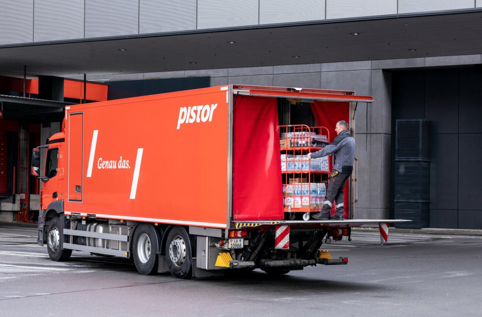 Un camion de livraison immatriculé Pistor est garé, porte arrière ouverte. Un employé décharge un chariot rempli de bouteilles de boissons devant un bâtiment moderne.