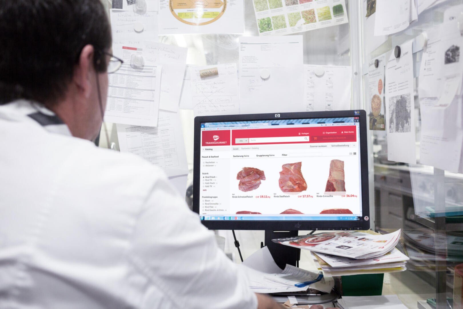 Une personne en blouse blanche est assise à un bureau encombré, regardant un écran d'ordinateur affichant différentes coupes de viande sur un site web. Des papiers et des notes sont épinglés au mur autour de l'espace de travail.