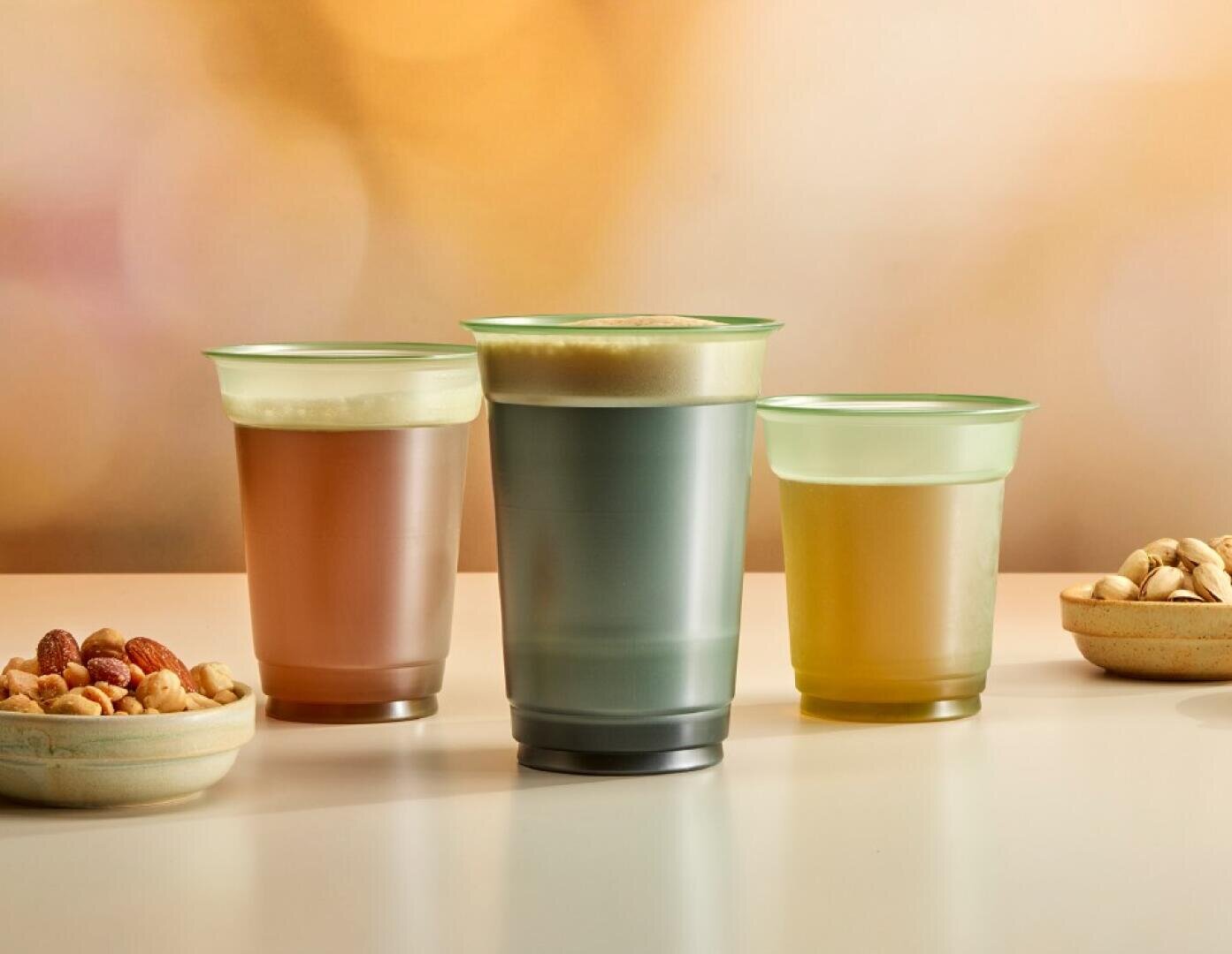 Trois boissons dans des gobelets en verre au lait, chacune avec une couche de mousse, sont posées sur une surface claire. Deux petits bols contenant différentes noix et pistaches sont posés à côté des gobelets. L'arrière-plan est doucement flou.