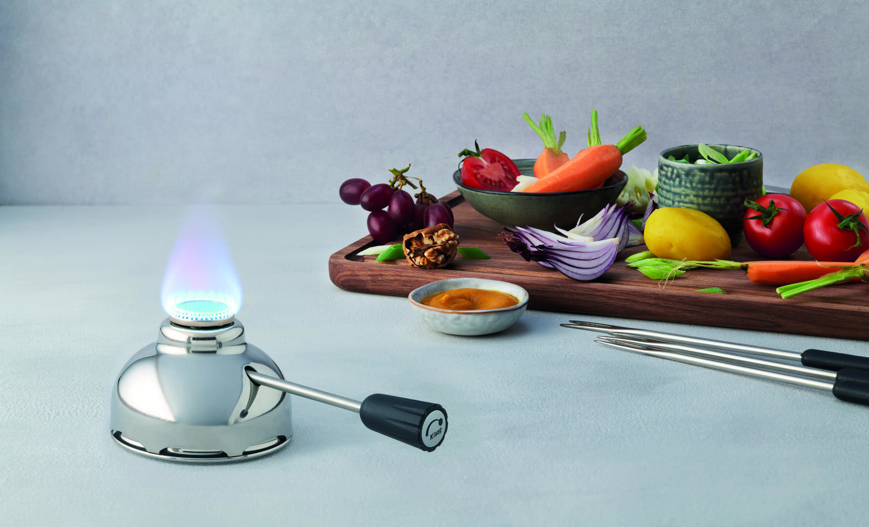 Auf einer Theke steht neben Fonduegabeln ein Küchenbrenner mit blauer Flamme, im Hintergrund ist ein Holzbrett mit frischem Gemüse, Trauben und Soße zu sehen.