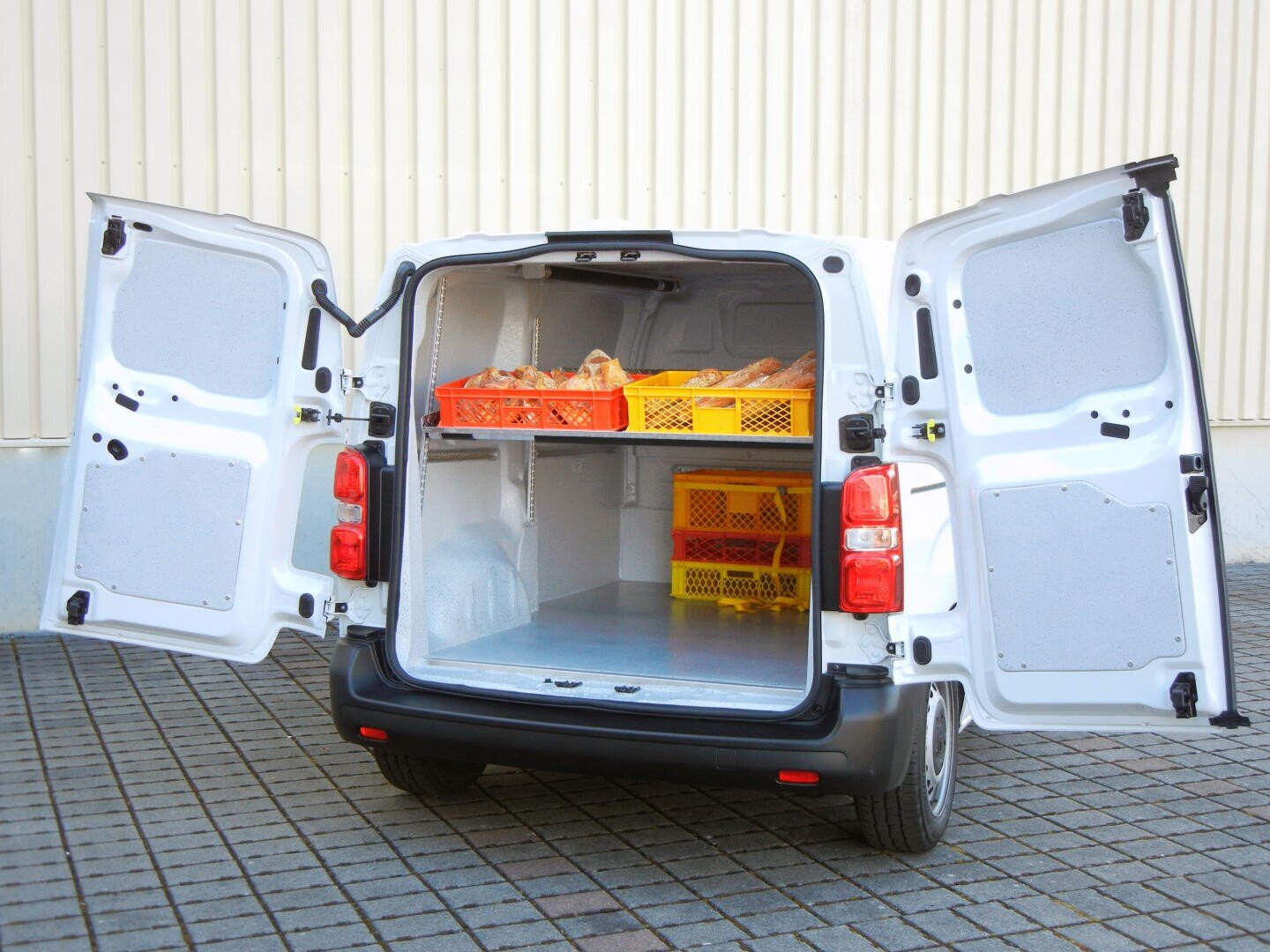 Ein weißer Lieferwagen mit geöffneten Hecktüren, in denen sich gelbe und orangefarbene Kisten mit Brot und Brötchen befinden. Der Lieferwagen ist auf einer gepflasterten Fläche neben einem beigen Gebäude geparkt.