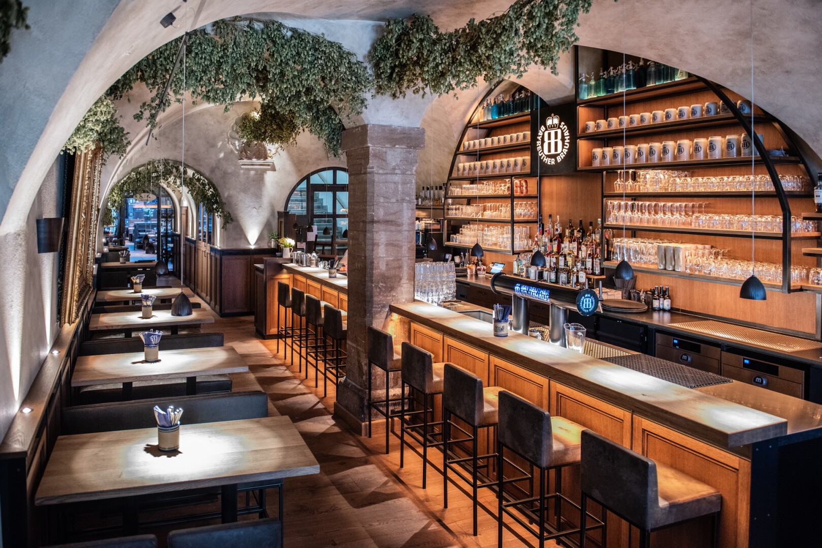 Eine stilvolle, moderne Bar mit gewölbten Decken, hängenden Pflanzen, Holztischen, hohen Barhockern und Regalen voller Gläser und Flaschen an der Rückwand, die eine warme und einladende Atmosphäre schaffen.