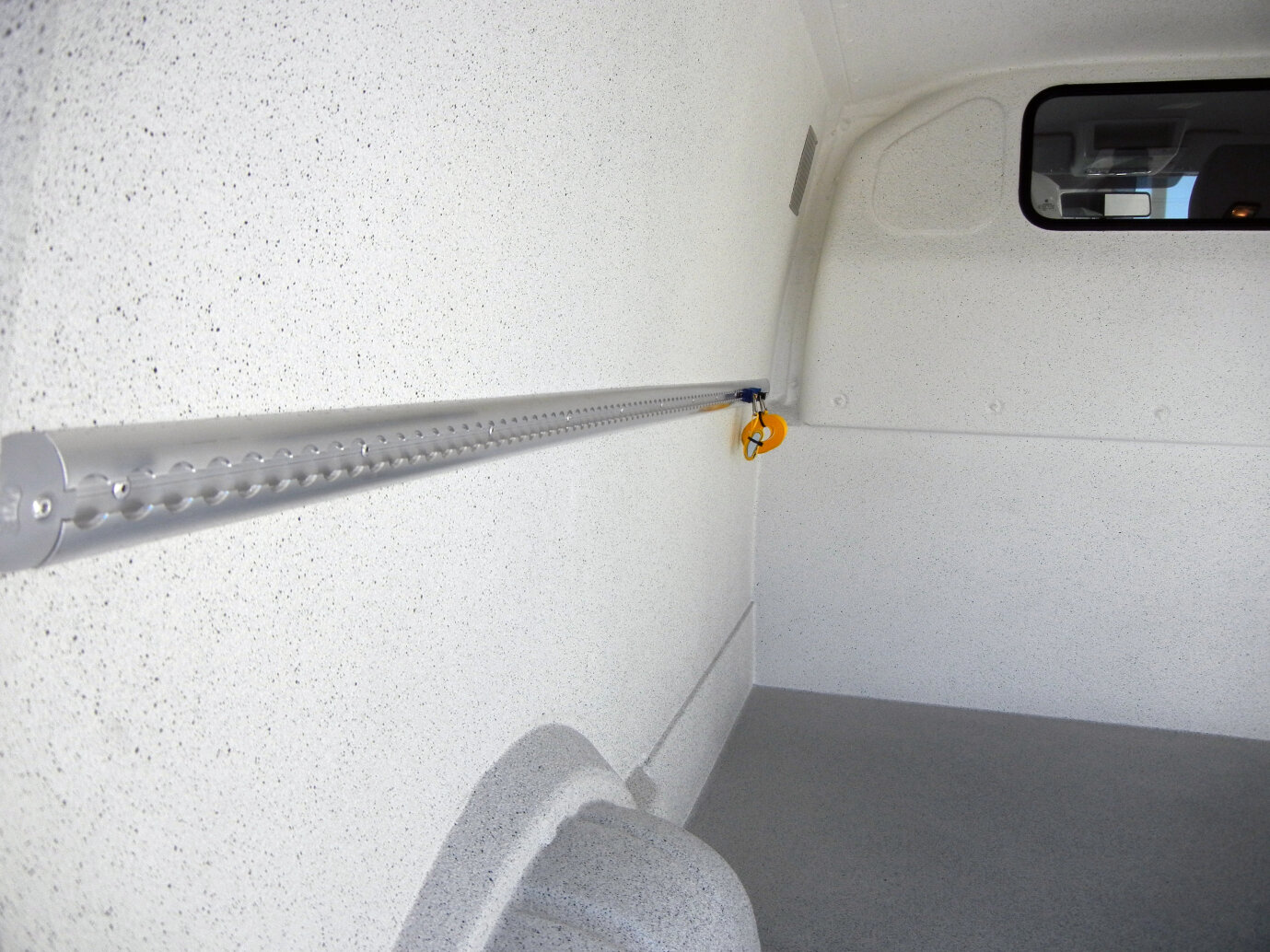 Das Innere eines weißen, leeren Lastwagens mit einer gesprenkelten Wand, einem grauen Boden, einer horizontalen Metallschiene auf der linken Seite und einem kleinen Fenster auf der rechten Seite nahe der Decke.